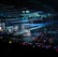 A large indoor arena filled with an audience watching a live performance. The stage features large screens displaying a performer and graphics, with bright spotlights and ambient lighting creating a dynamic atmosphere. The crowd is holding glow sticks, adding vibrant colors to the scene.