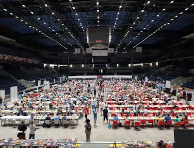 An indoor sports arena filled with rows of tables, each occupied by people engaged in competitive activities. The ceiling is high, with numerous lights illuminating the large space. The audience seating on either side is empty, and there are flags hanging from the upper level.