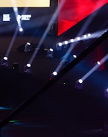 An array of bright stage lights beams across a dark arena, creating a dynamic atmosphere with a large screen displaying various logos and text. A camera crane is visible in the foreground, suggesting a professional event setup.
