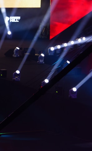 An array of bright stage lights beams across a dark arena, creating a dynamic atmosphere with a large screen displaying various logos and text. A camera crane is visible in the foreground, suggesting a professional event setup.