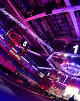 An indoor arena with vibrant purple and blue lighting, featuring large numbered screens suspended from the ceiling. The venue has a high ceiling with intricate metal truss structures and hanging lights, creating a futuristic and energetic atmosphere.